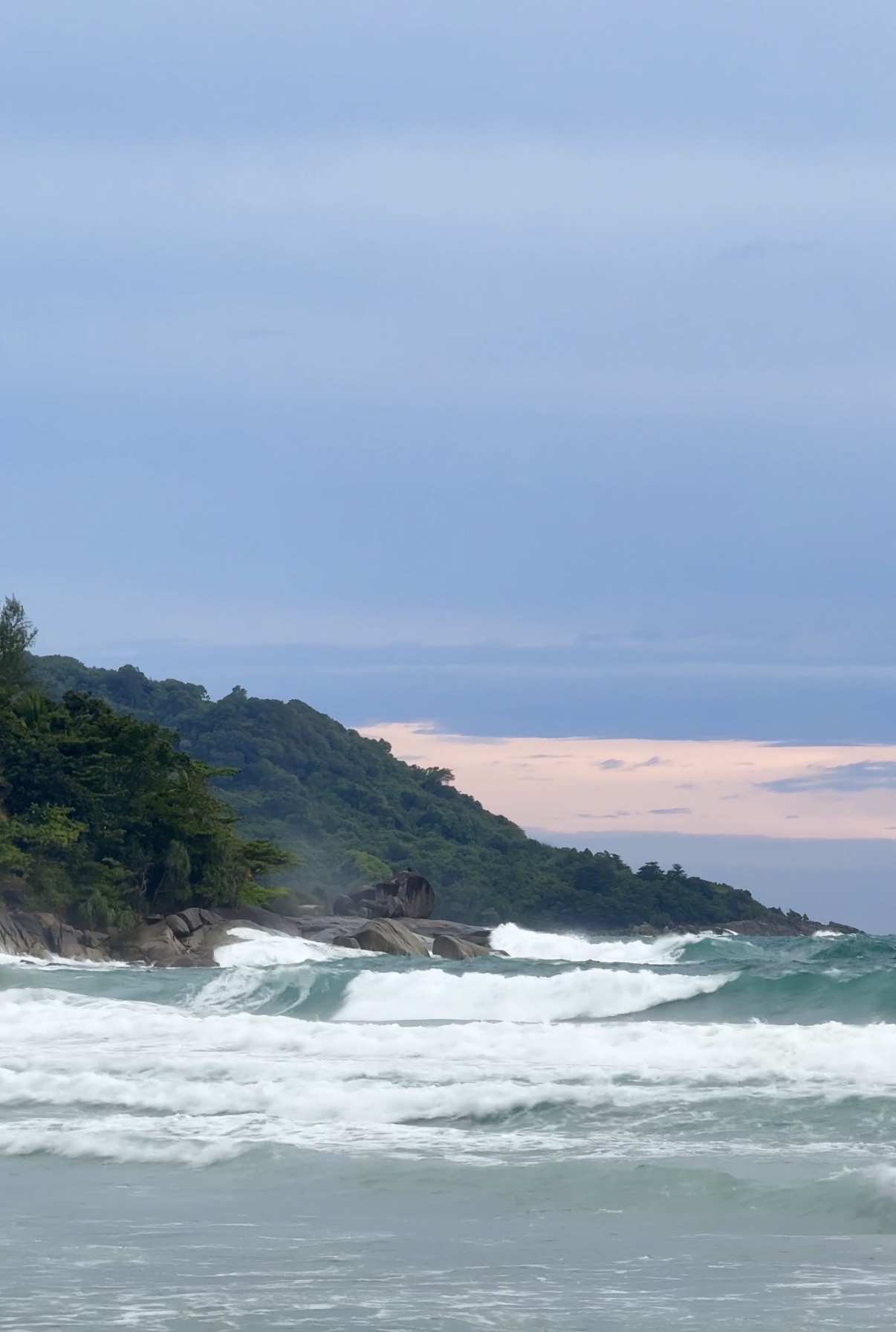 Waves breaking beside a wooded headland under an overcast sky in Phuket